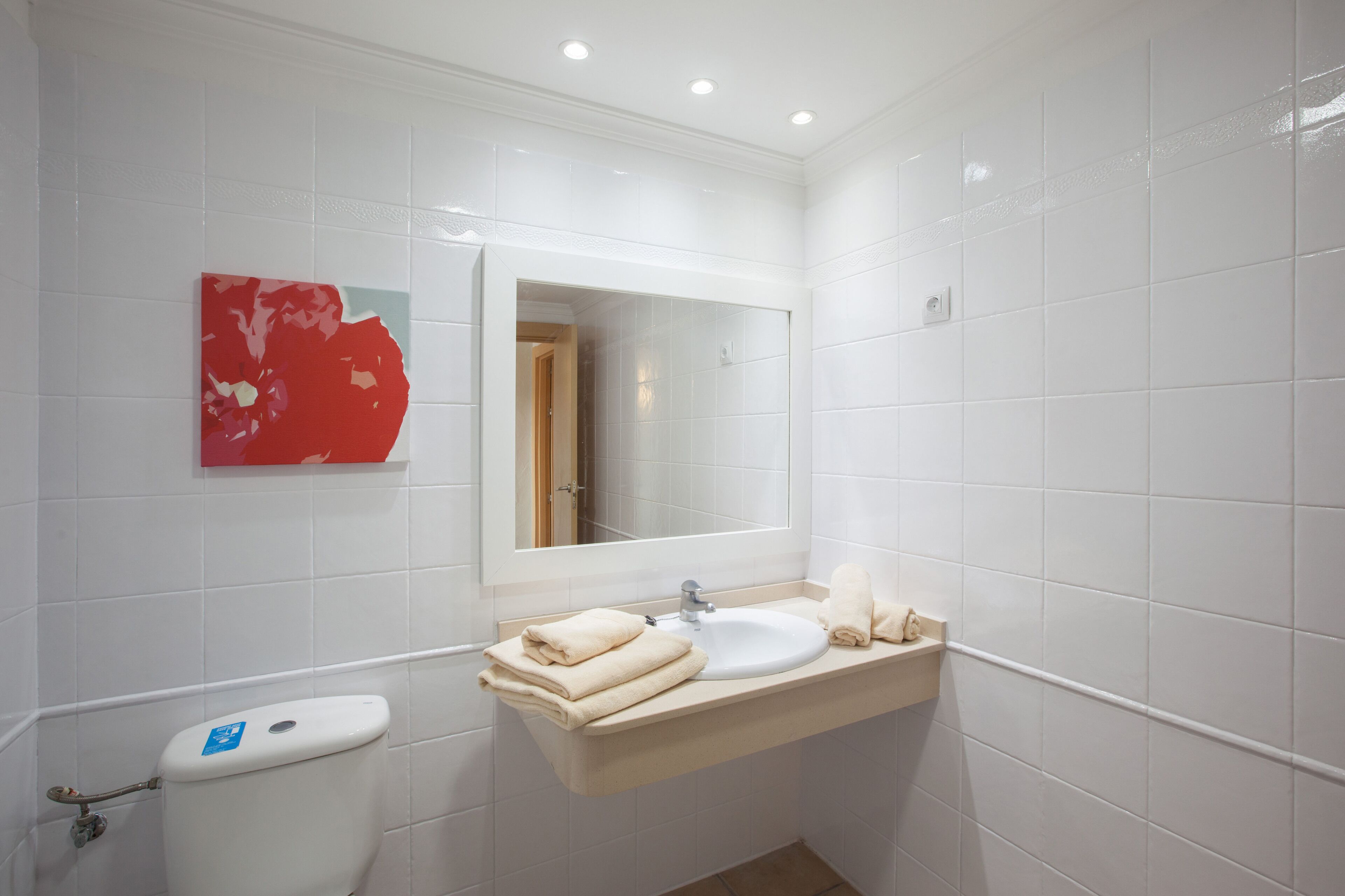 Un baño con azulejos blancos, un lavamanos, un inodoro y un cuadro abstracto rojo en la pared.