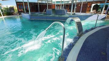 Una gran piscina interior con agua cristalina, zona de jacuzzi y área de descanso cubierta con sillones reclinables.