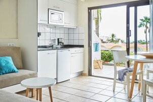 Una cocina moderna con armarios blancos, microondas y cafetera. Hay un comedor con una mesa redonda y sillas. Una puerta corrediza de vidrio da a un balcón con vistas a palmeras y un edificio.