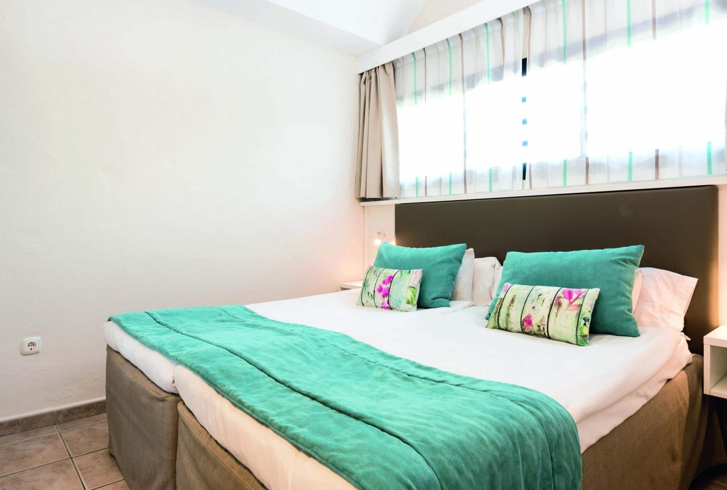 A bedroom with a bed, a headboard, a window with curtains, and a bedside table.
