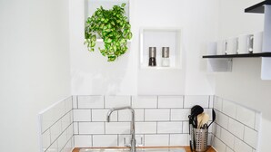Una cocina con pileta de acero inoxidable, salpicadero de azulejos blancos, encimera de madera, una planta colgante, un especiero y un portalapices.