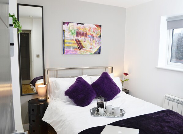 A modern hotel room with a neatly made bed, a coffee table with a tray, a painting on the wall, and a window with a view.