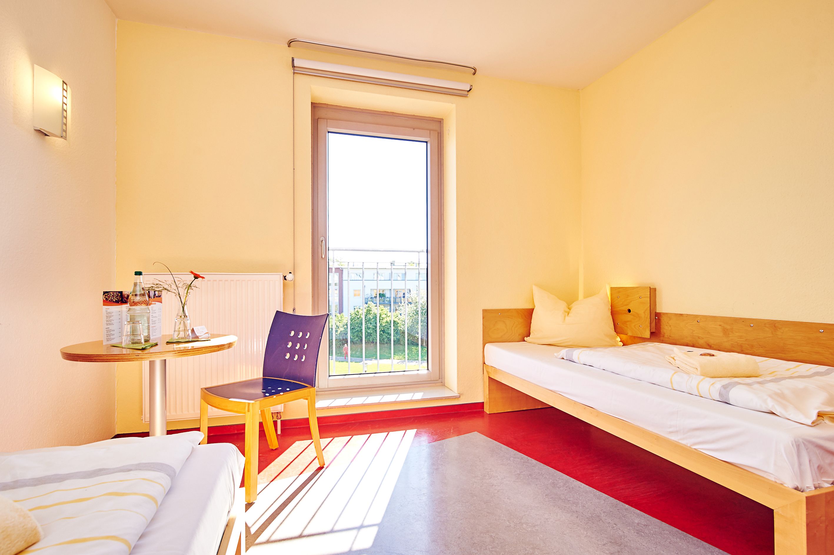 A hotel room with a bed, a chair, a small table with a bottle and a vase, and a window with a view of buildings.
