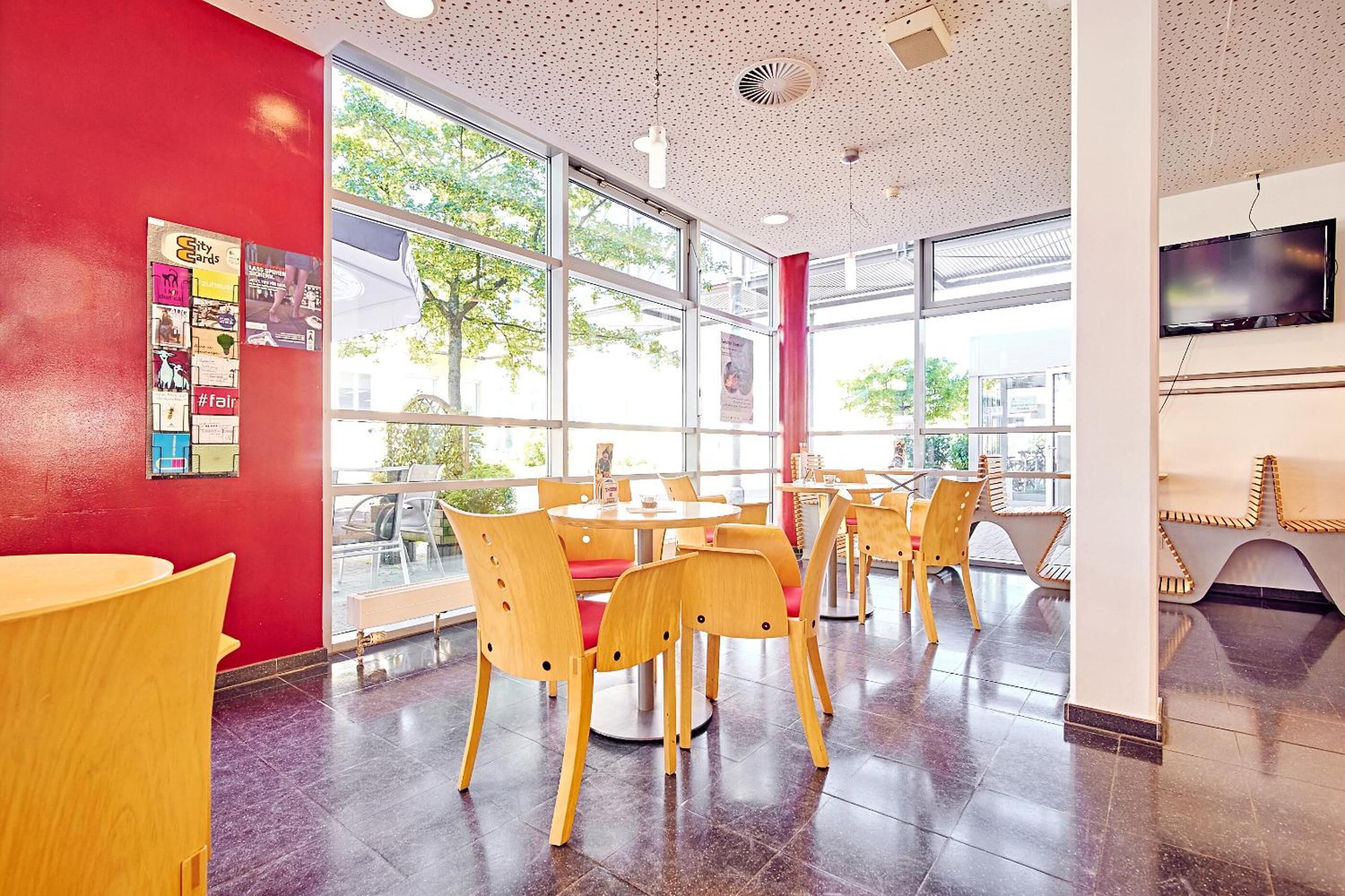 A modern interior with round tables and wooden chairs, large windows, and a red accent wall.