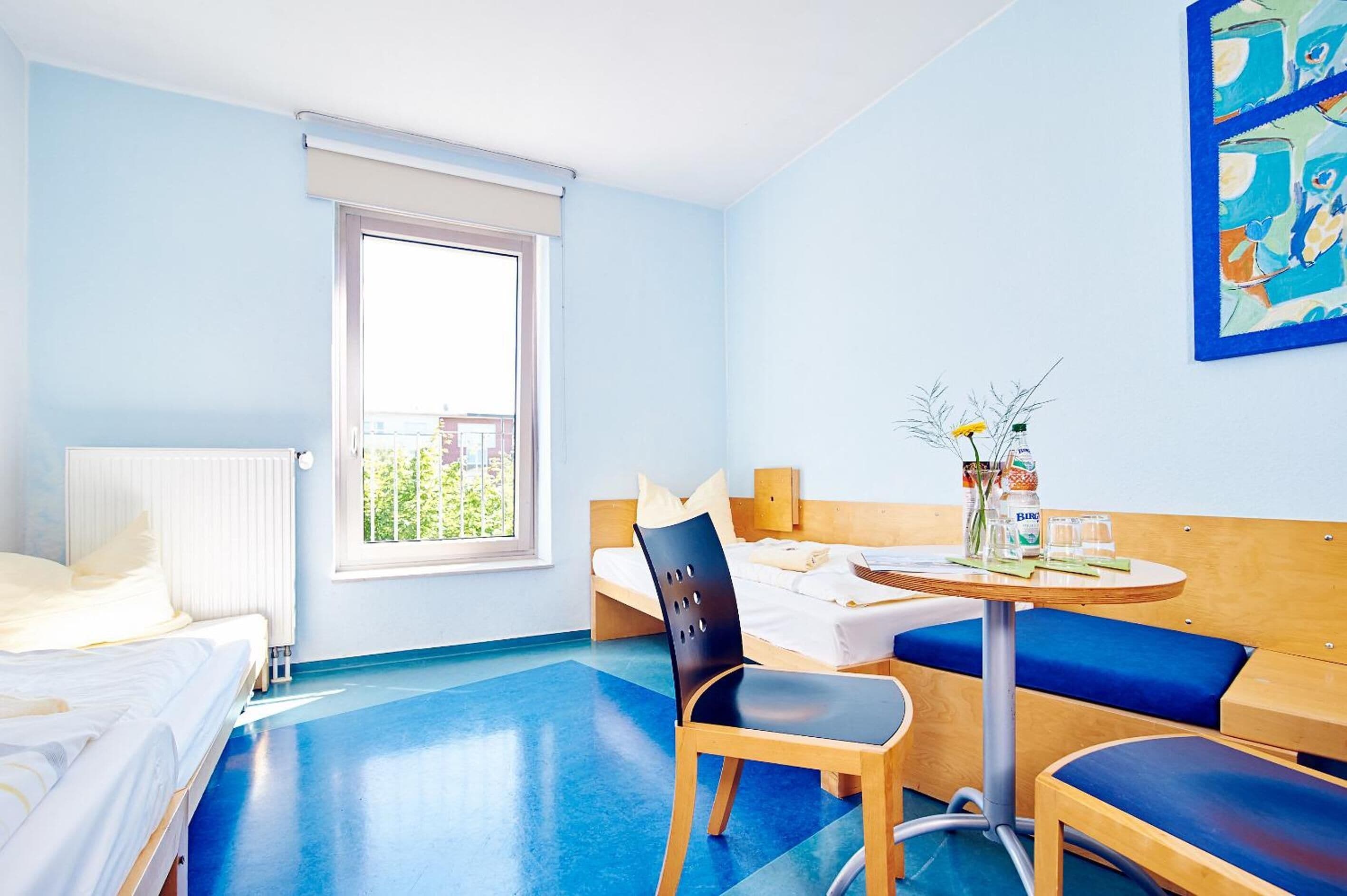 A hotel room with a bed, a chair, a small table with a vase and bottles, and a window with a view of greenery.