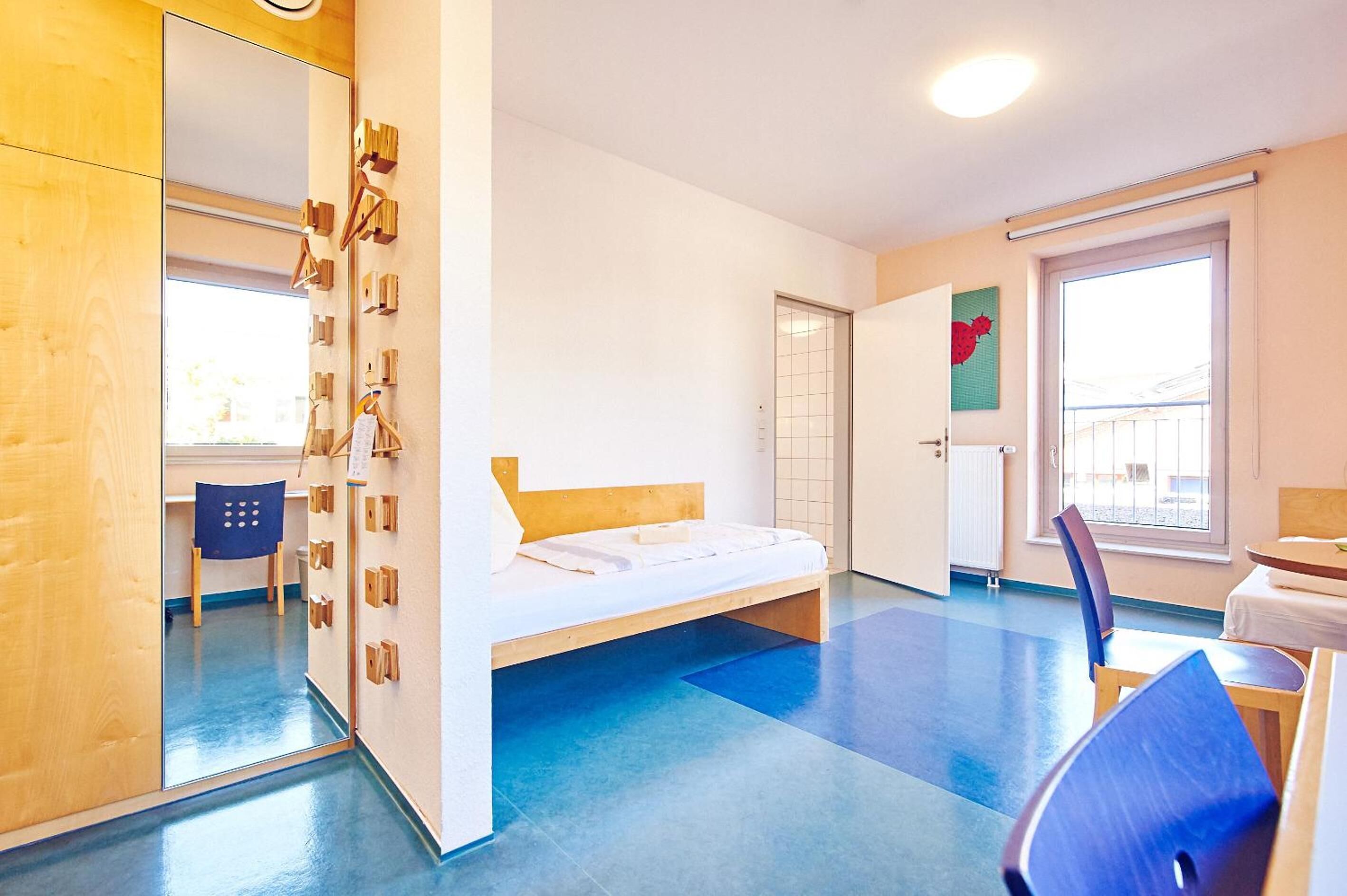 A modern hotel room with a single bed, a desk, and a chair. There is a large window providing natural light and a view of the outside.