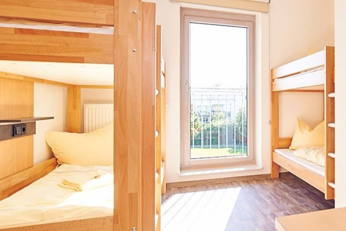 A compact hotel room with two bunk beds, a small kitchenette, a sliding glass door to a balcony, and a small table with a vase of flowers.
