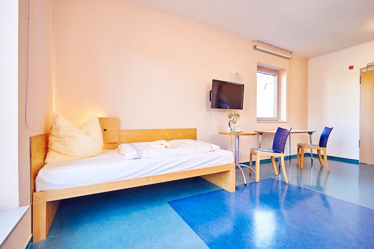 A hotel room with a bed, a TV mounted on the wall, a small table with a chair, and a window providing natural light.