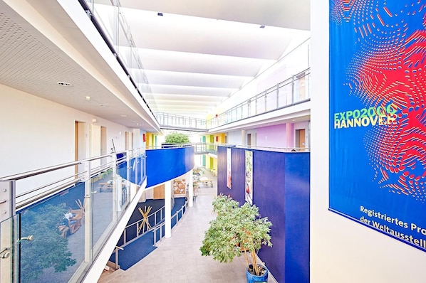 A modern indoor corridor with glass railings, a central staircase, and a banner for Expo 2020 Hannover.
