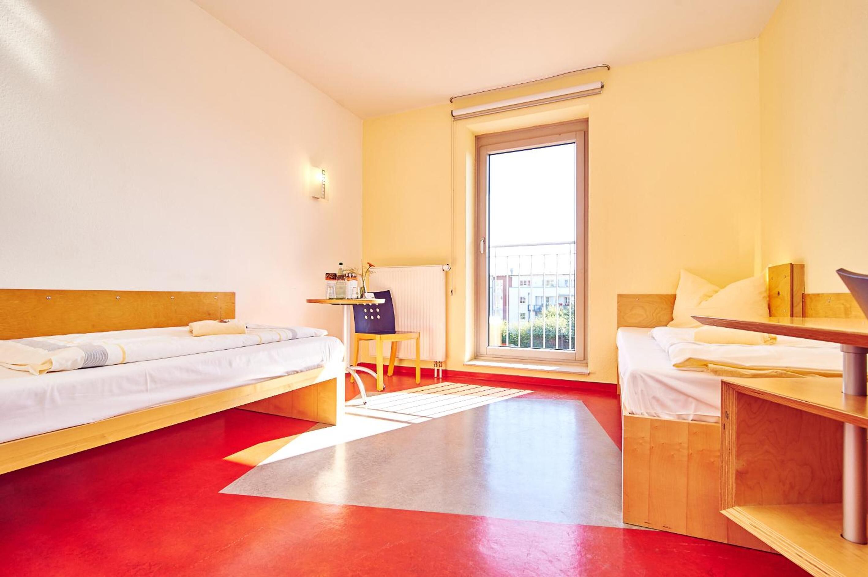 A hotel room with two beds, a small table, a chair, a radiator, and a window with a view of buildings.