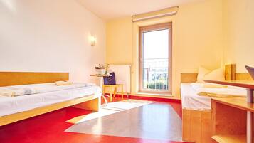 A hotel room with two beds, a small table, a chair, a radiator, and a window with a view of buildings.