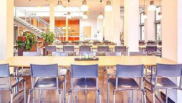 Una amplia cafetería con numerosas mesas y sillas, una escalera al fondo y plantas en macetas.
