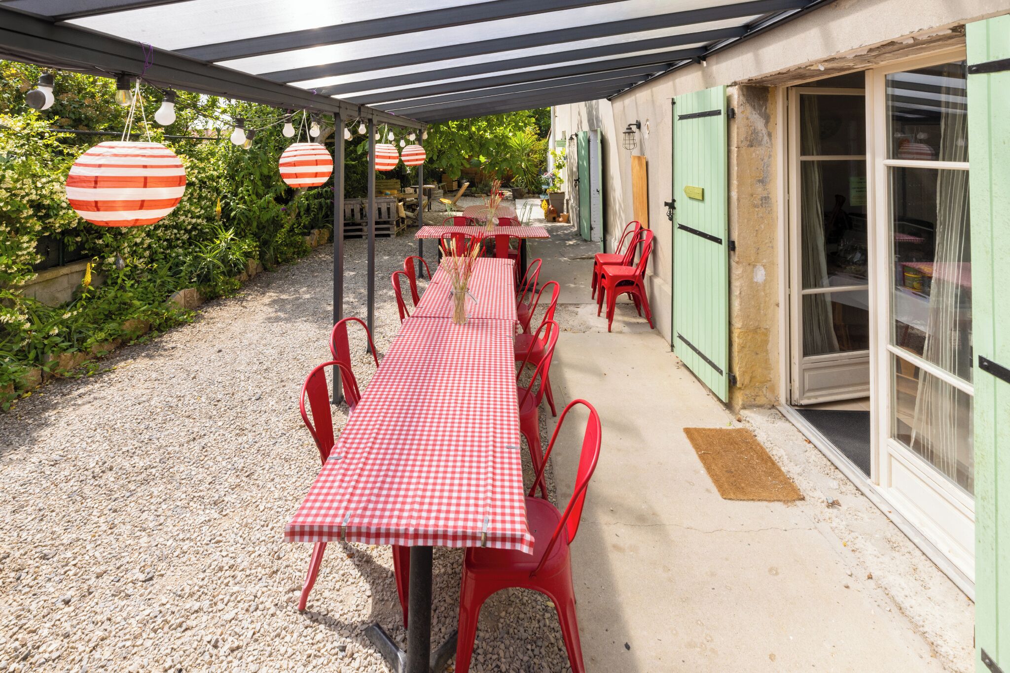 Un patio al aire libre con una mesa de cuadros y sillas rojas, rodeado de vegetación y un camino de grava.