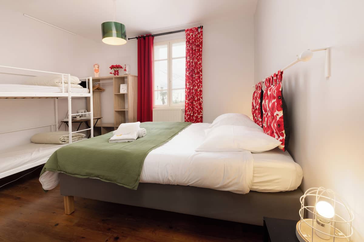 A bedroom with a bunk bed, a single bed, a green blanket, a white pillow, a red curtain, a window, and a bedside lamp.