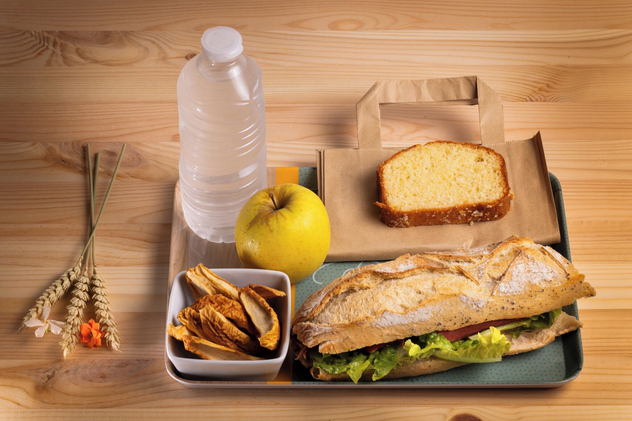 Una bandeja con un sándwich, una botella de agua, una manzana y algunos bocadillos sobre una mesa de madera.