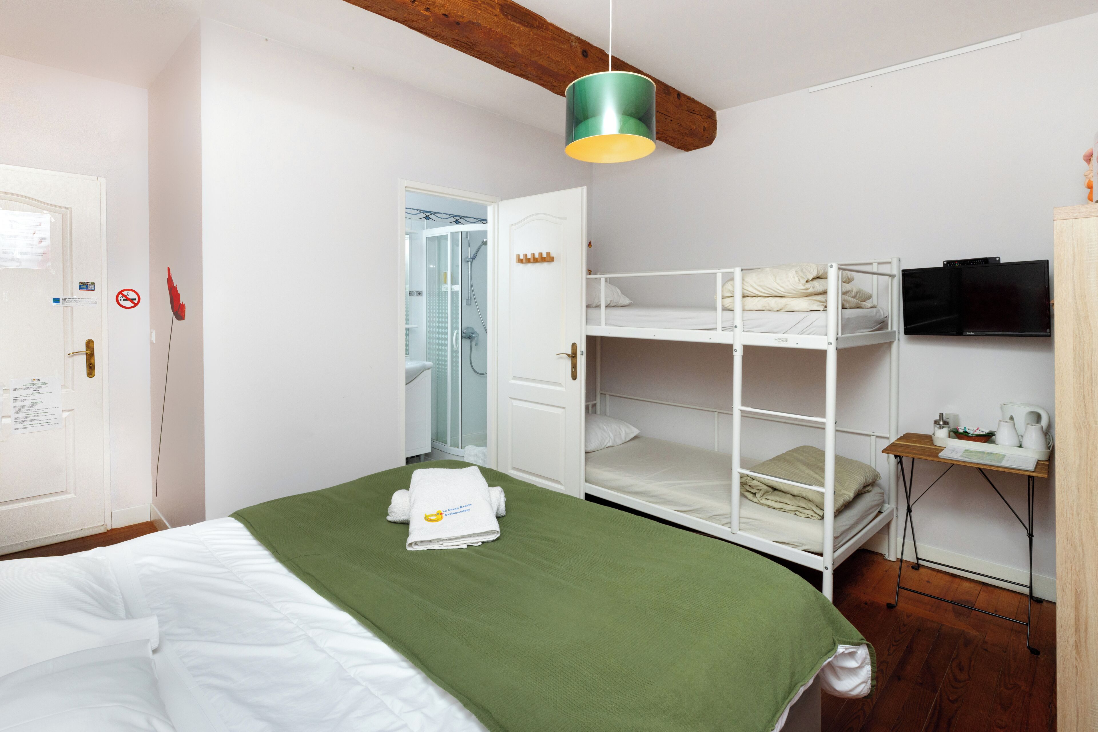Una camera da letto con un letto a castello, una coperta verde, una porta bianca, una trave in legno al soffitto, una televisione e un tavolino con un bollitore.