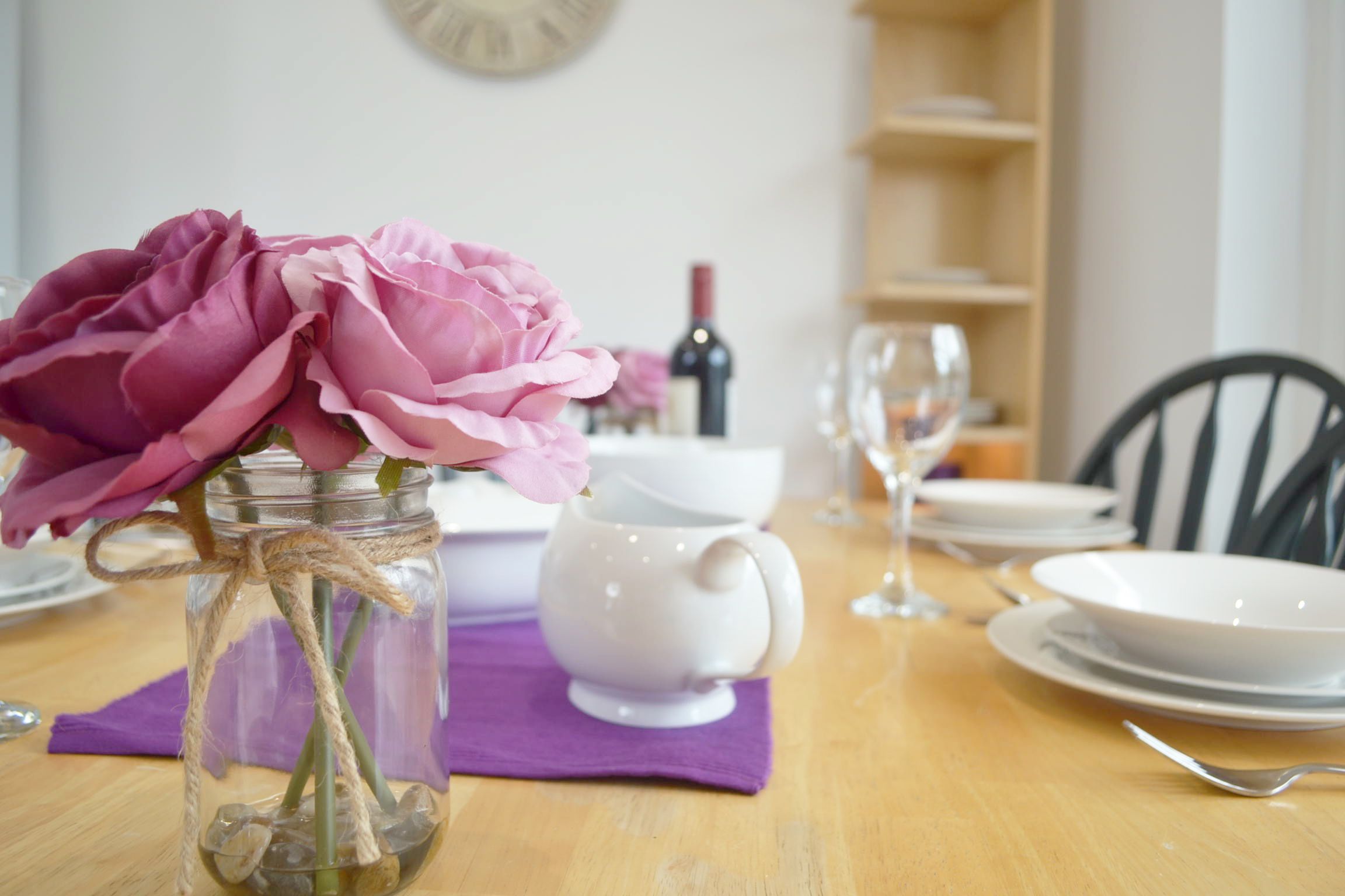 Een eettafel met een vaas roze bloemen, een witte karaf en een wijnfles op de achtergrond.