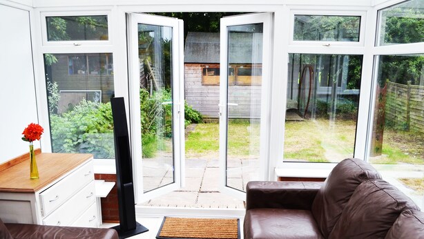 A modern living room with a brown leather sofa, a wooden coffee table, and a TV on a stand. Large glass doors lead to a garden with a wooden deck and a small shed. - No 40, 5 beds, sleeps 7 (Coventry)
