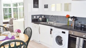 A modern kitchen with a dishwasher, washing machine, and oven. There is a dining area with a table and chairs, and a window with a view of greenery. - No 40, 5 beds, sleeps 7 (Coventry)