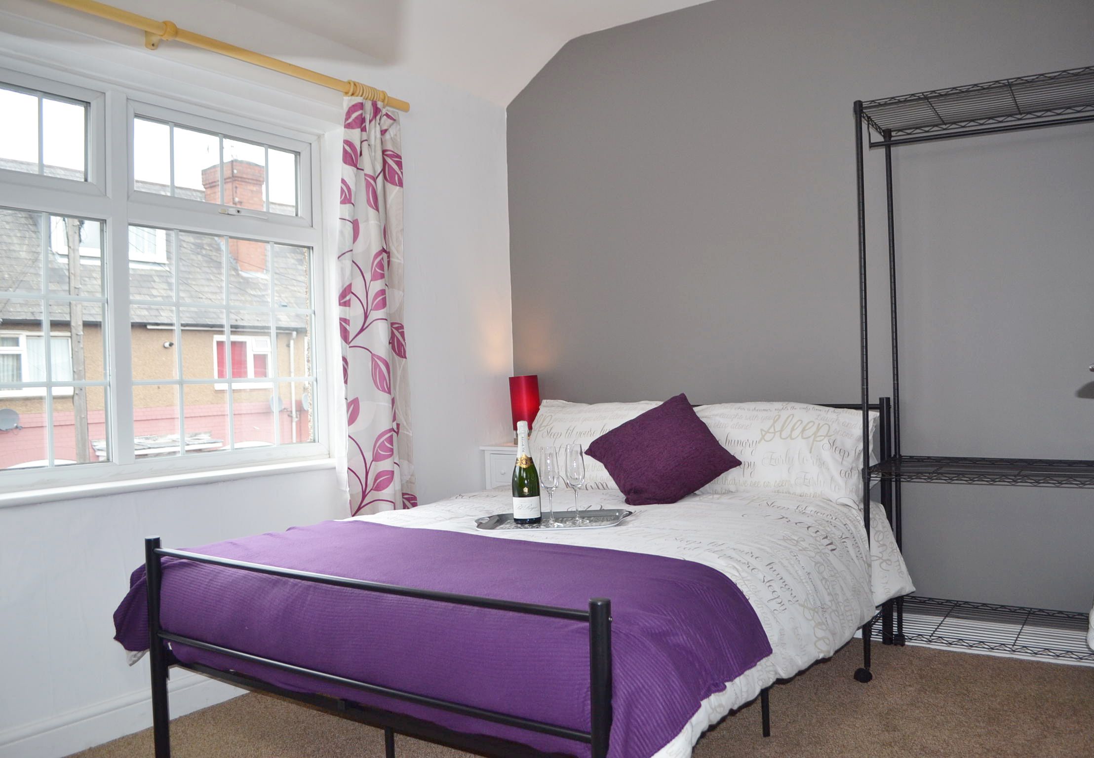 A bedroom with a bed, a window with curtains, a bedside table with a bottle and glasses, and a metal shelf unit.