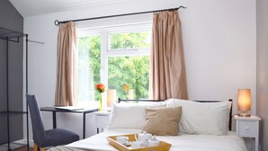 A neatly made bed with a tray of white cups and a teapot, a bedside table with a lamp, and a chair in a room with a window and curtains. - No 40, 5 beds, sleeps 7 (Coventry)