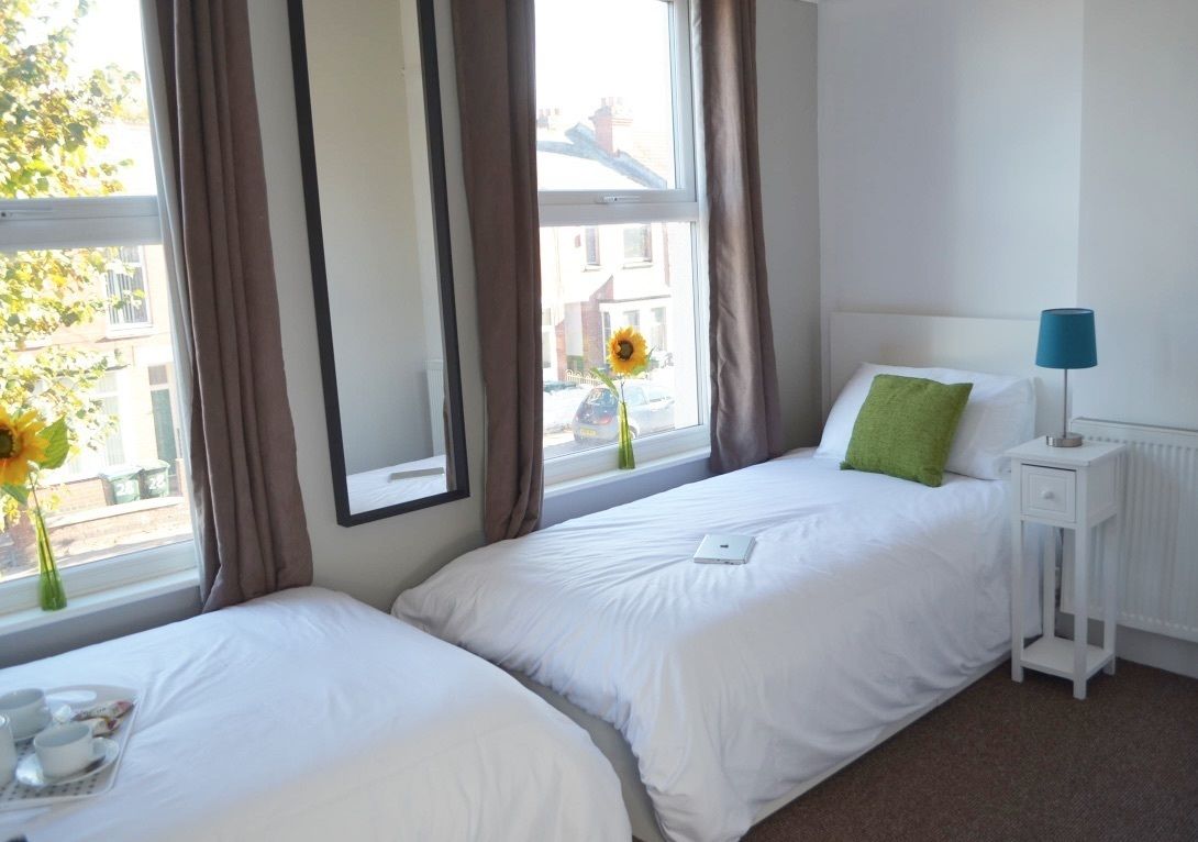 A hotel room with two beds, a window with curtains, a bedside table with a lamp, and a radiator.