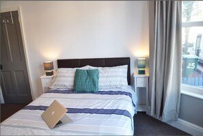 A neatly made bed with a laptop on it, two bedside tables with lamps, a window with curtains, and a door. - TwentyThree (Coventry)