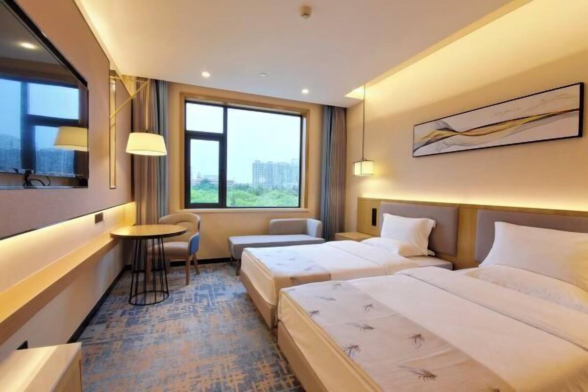 A hotel room with two beds, a small table, a chair, a window with a view, and a painting on the wall.