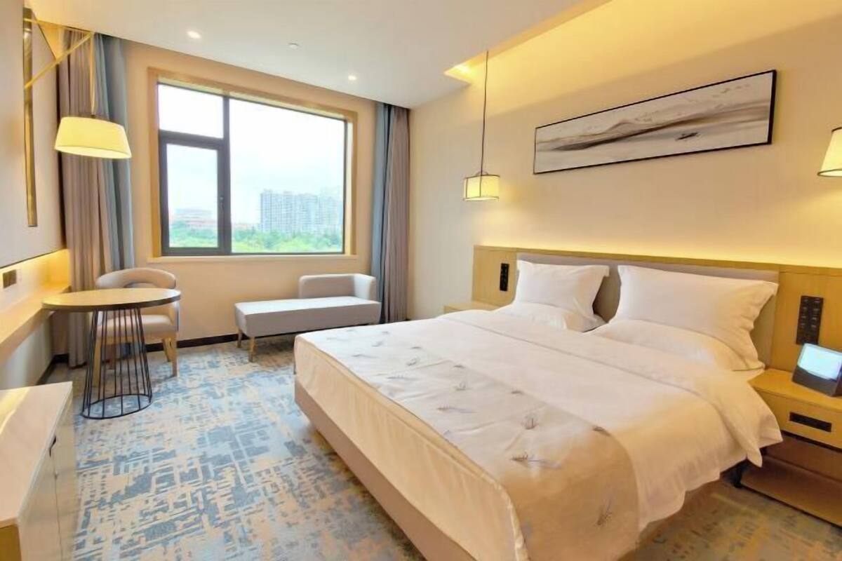 A hotel room with a bed, a desk, a chair, a window with a view, and a painting on the wall.