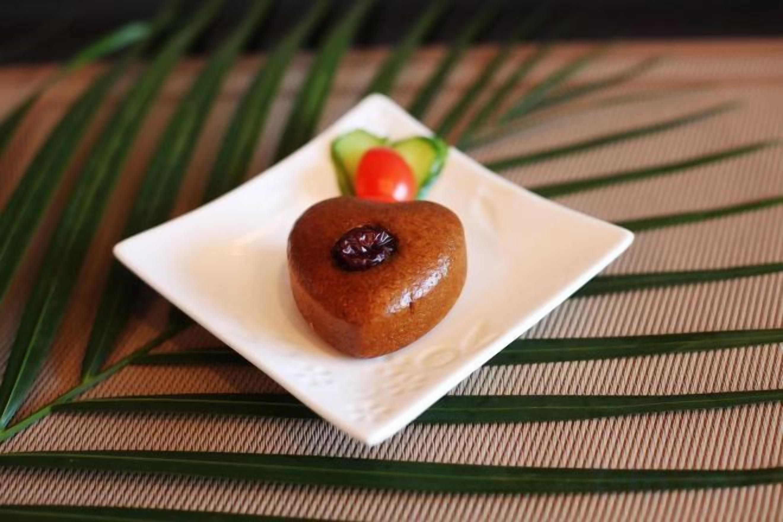 Postre sobre un plato cuadrado con una parte superior en forma de corazón, acompañado de una porción de aguacate y una cereza sobre una superficie texturizada.
