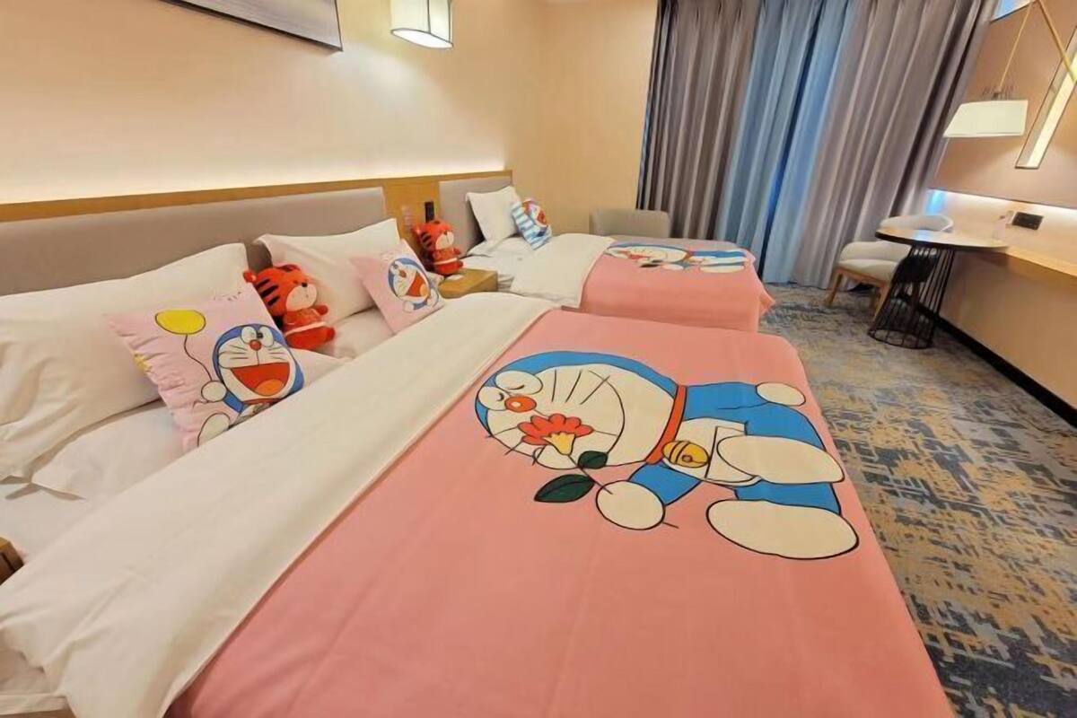 A hotel room with two beds, each with a cartoon character design on the bedspread. There is a desk with a chair and a lamp, and a window with curtains.