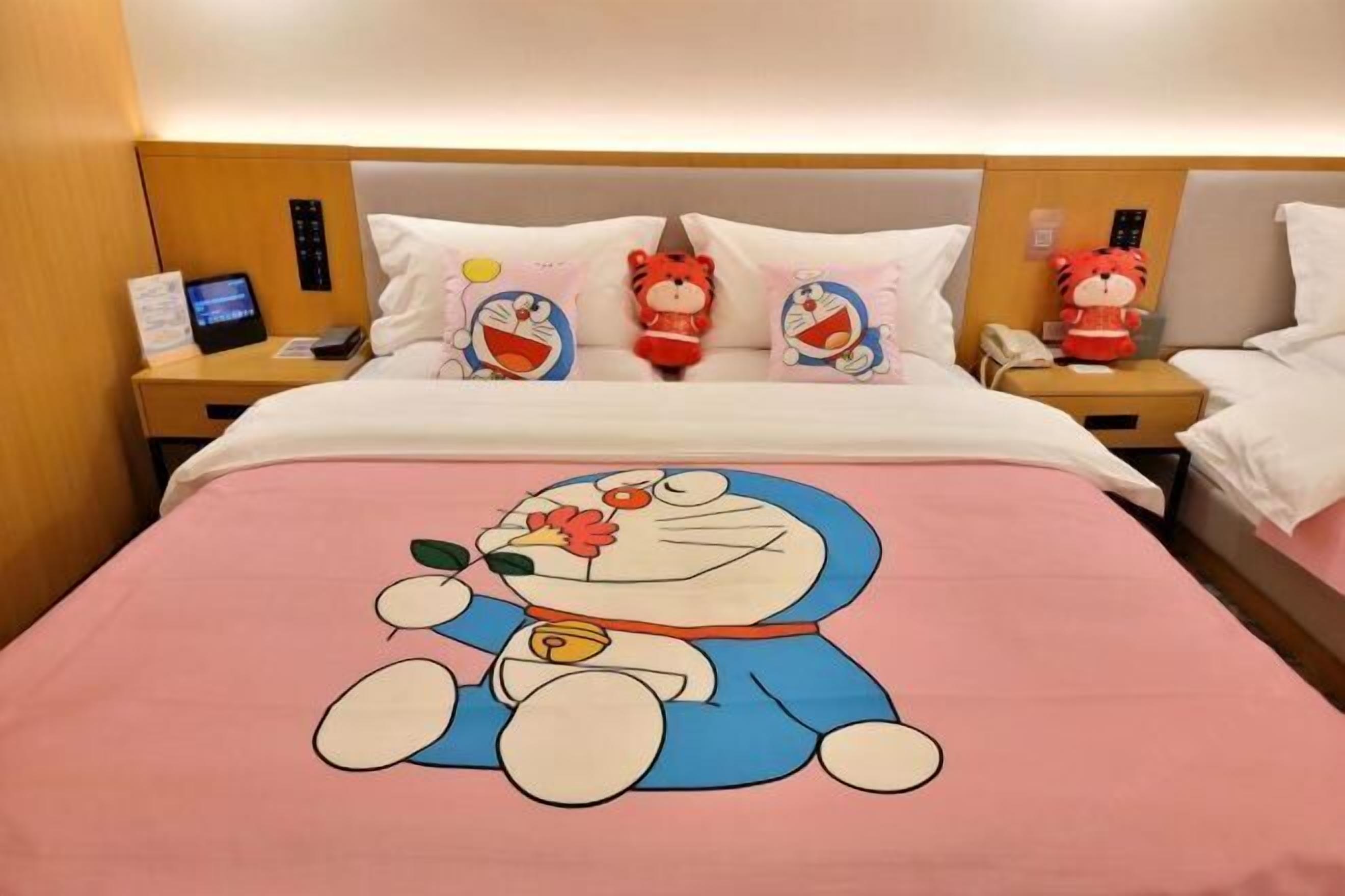 Une chambre d’hôtel avec un lit orné d’un motif représentant un personnage de dessin animé, deux tables de chevet équipées de téléphones et un écran numérique.