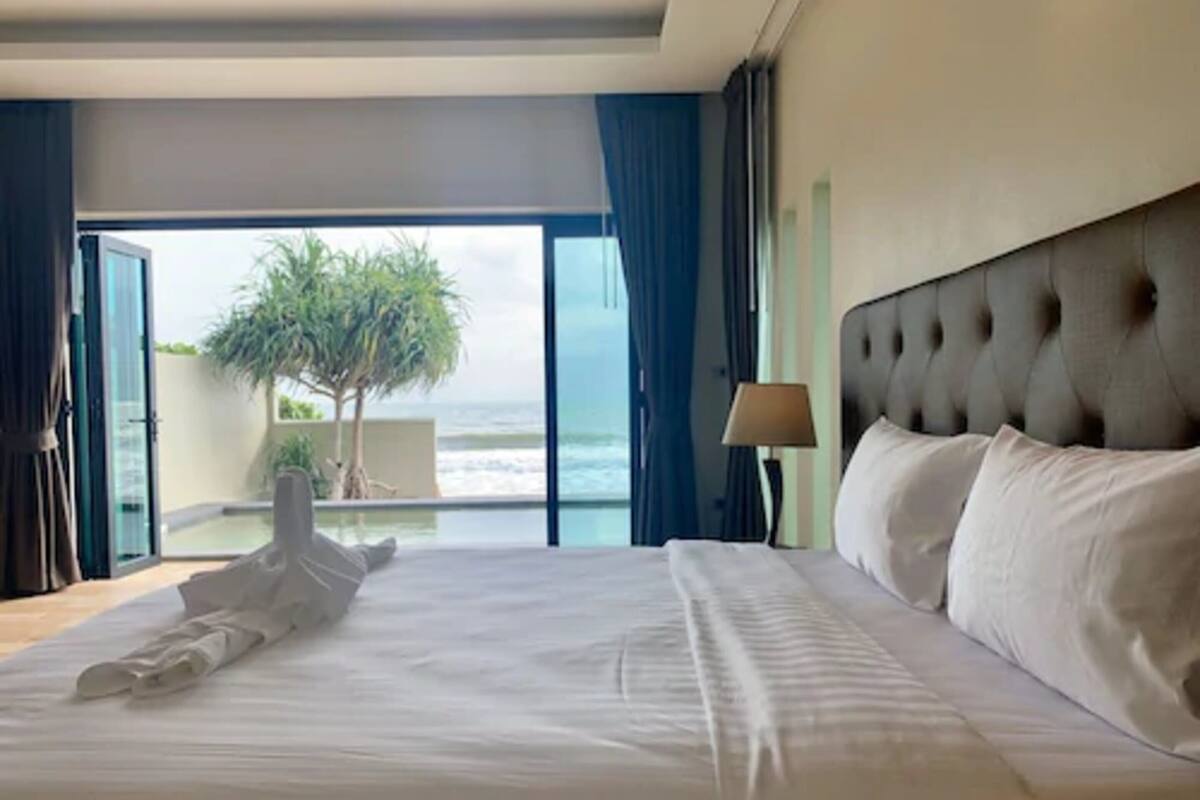 A hotel room with a large bed, a view of the ocean, and a balcony with a tree.