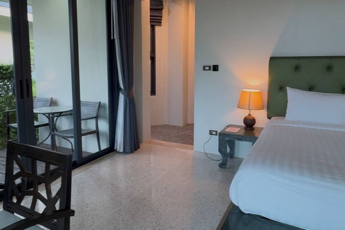 A hotel room with a bed, a nightstand, a lamp, a chair, and a view of the outdoors through a glass door.