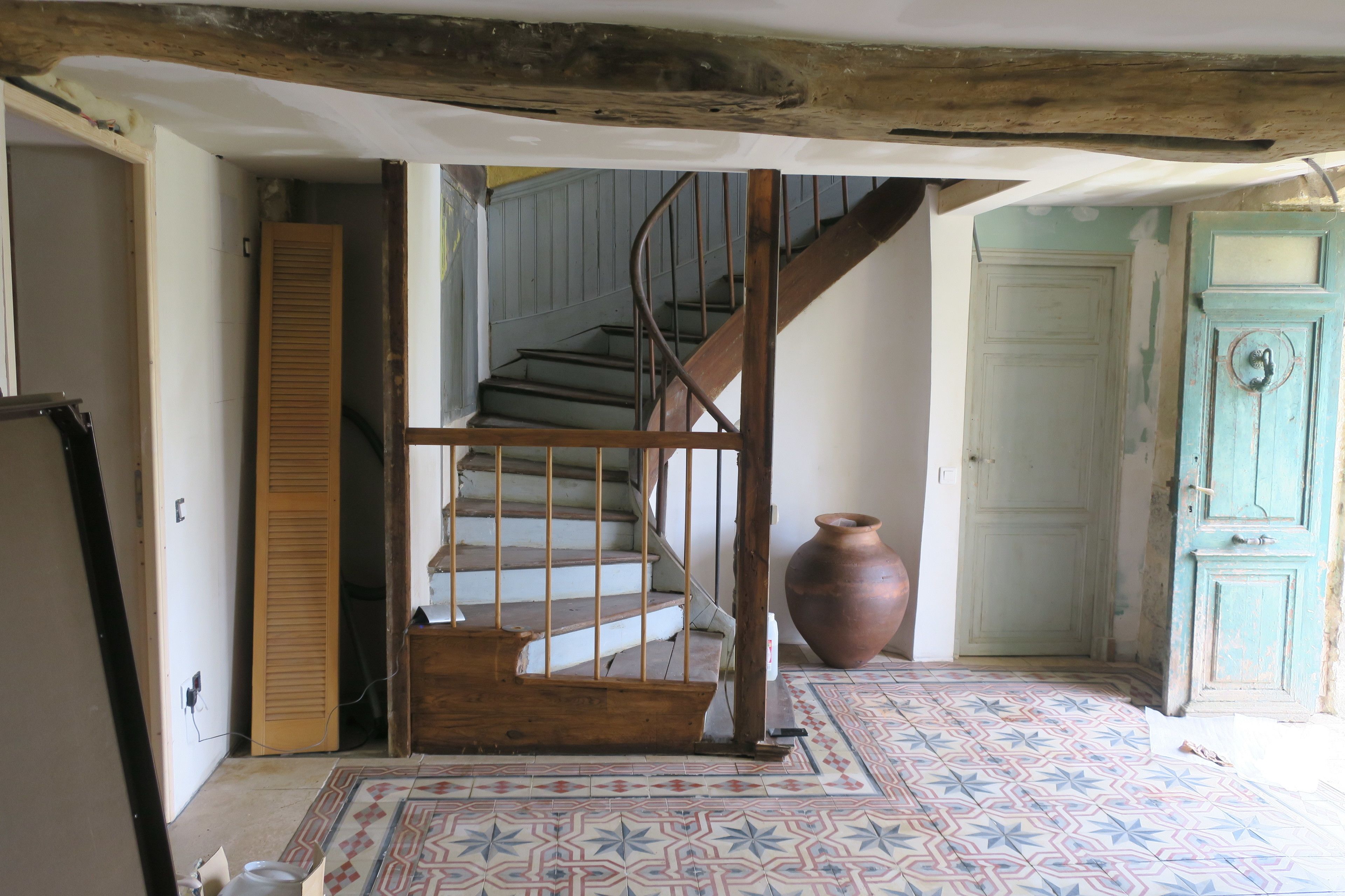 Une pièce avec un escalier en bois, un parquet à motifs, un grand vase et une porte au style rustique.