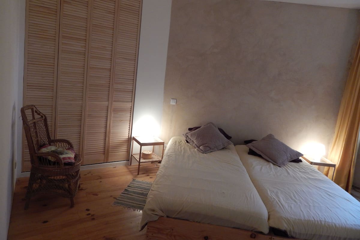 Un dormitorio con cama, dos mesitas de noche, una silla, un armario y una pequeña mesa con lámpara.