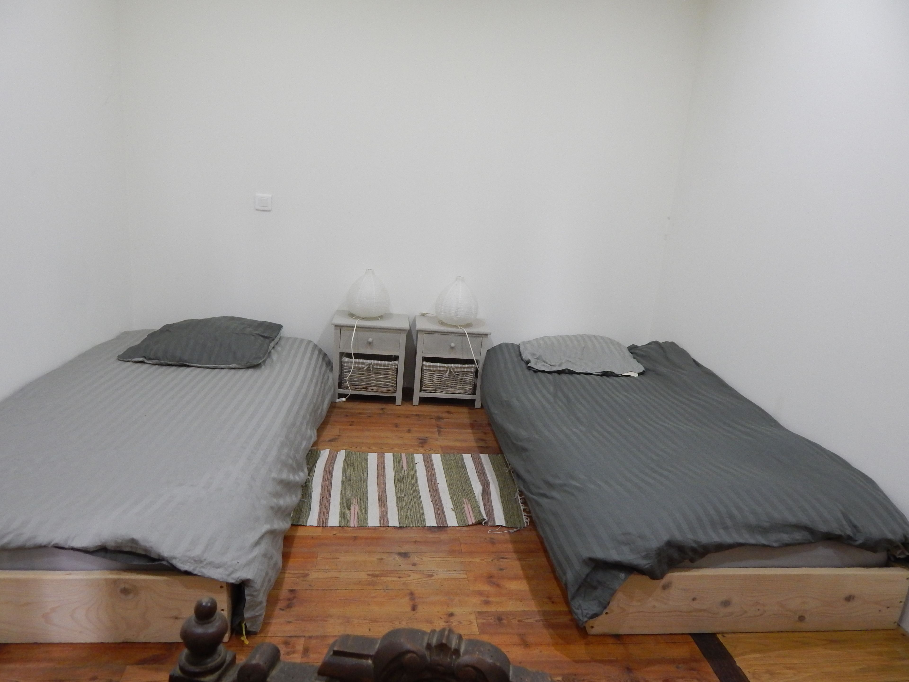 A small, simple room with two single beds, a small table with two lamps, and a woven rug on the wooden floor.