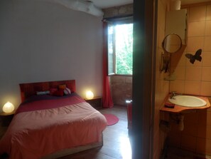 Een slaapkamer met een bed, nachtlampjes, een raam met gordijnen en een badkamer met een wastafel en spiegel.
