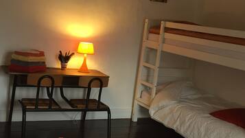 A dormitory room with a bunk bed, a desk with a lamp, a chair, and a patterned rug.