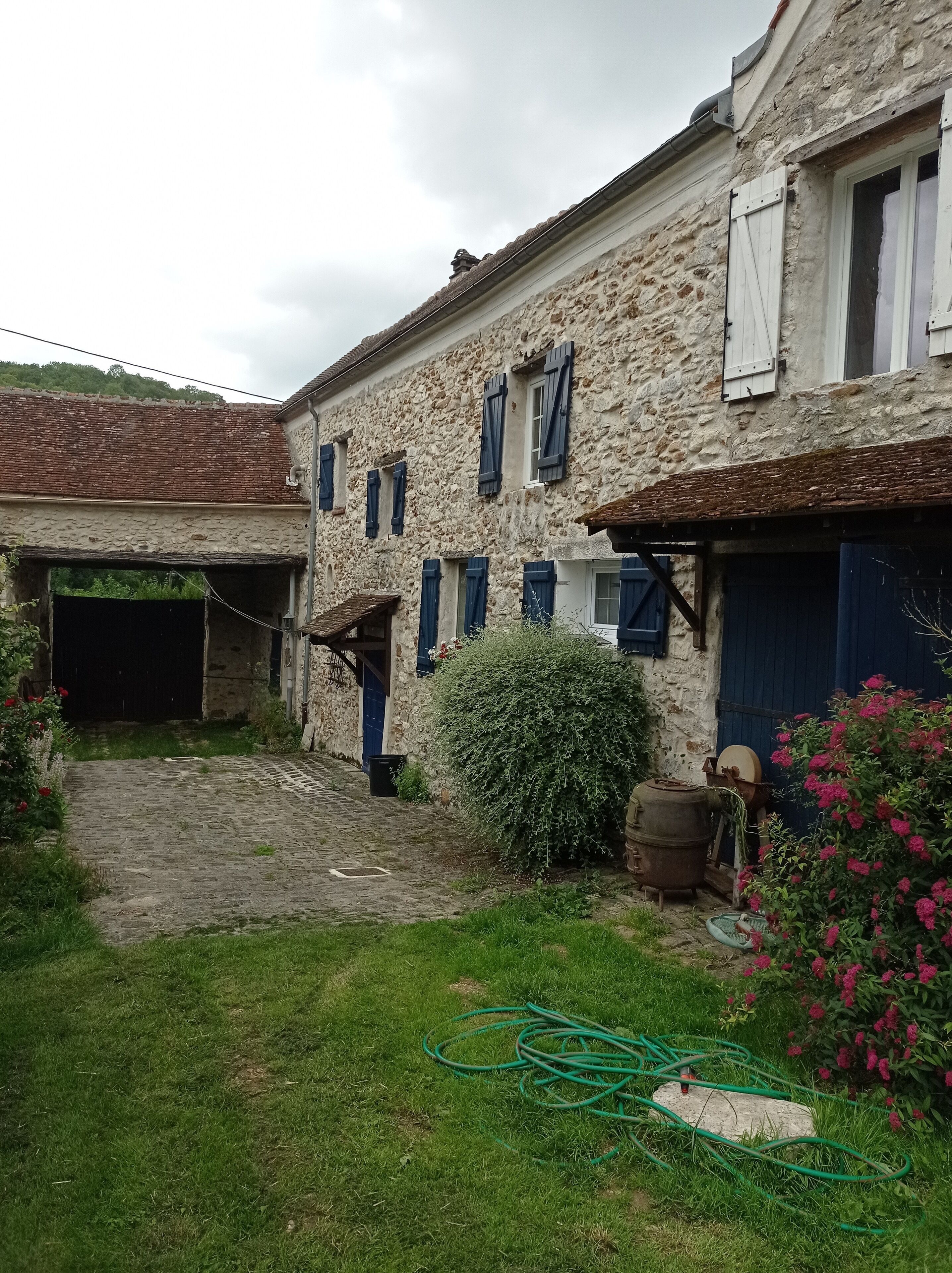 A stone building with multiple windows and doors, a cobblestone path leading to a garage, and a garden area with a hose and flowers.