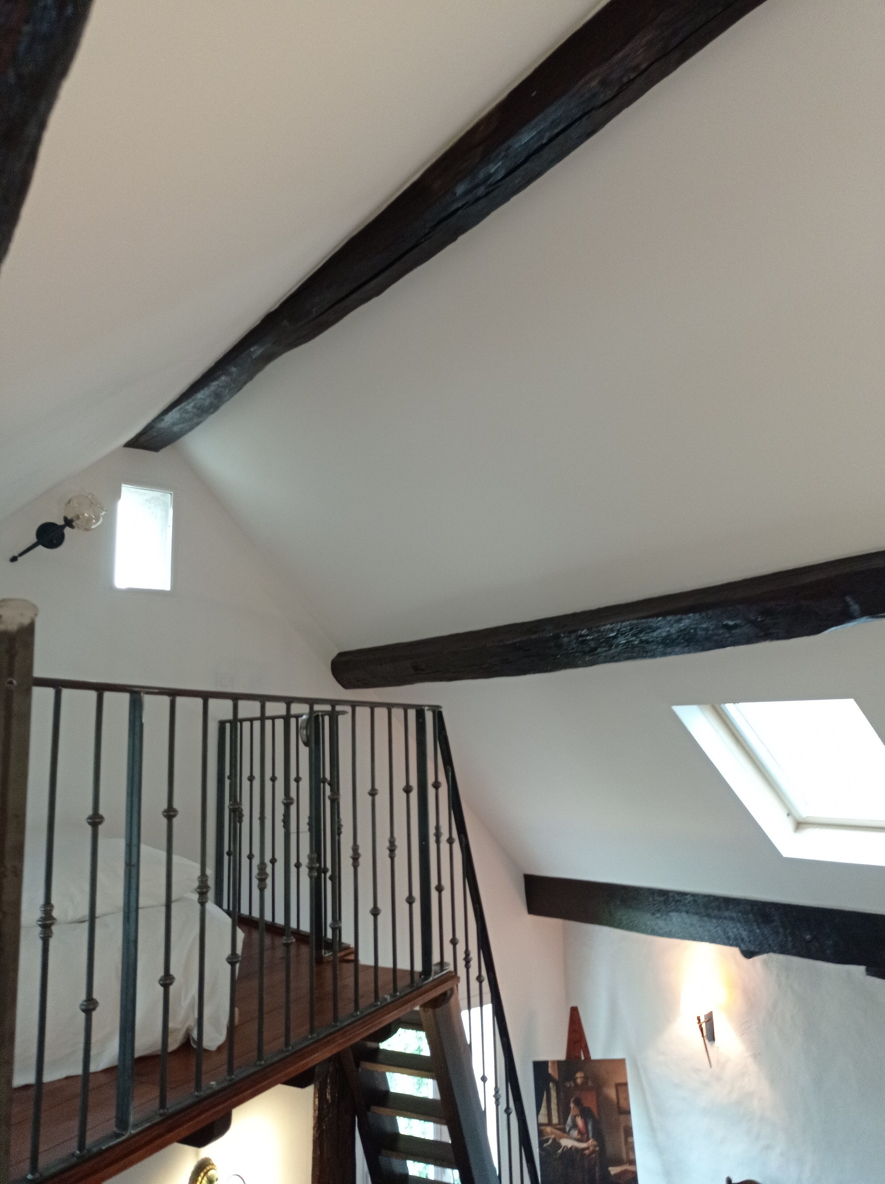 Eine Treppe mit hölzernem Geländer und Metallspindeln, die zu einer oberen Etage führt. Im Deckenbereich ist ein Oberlicht zu sehen, und ein Gemälde hängt an der Wand.