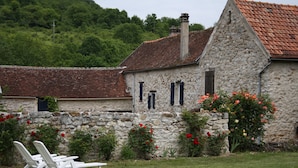 Un bâtiment en pierre avec un toit recouvert de tuiles rouges, un mur en pierre et un jardin agrémenté de fleurs rouges et de chaises blanches.