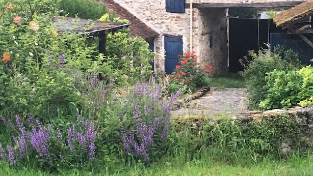 Un bâtiment en pierre doté d’un toit en pente, entouré d’une végétation luxuriante et de plantes en fleurs.