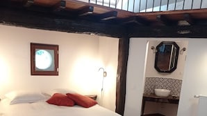 Une chambre avec un plafond en bois, un lit avec une literie blanche, un oreiller rouge, un miroir fixé au mur et une fenêtre donnant sur le ciel.