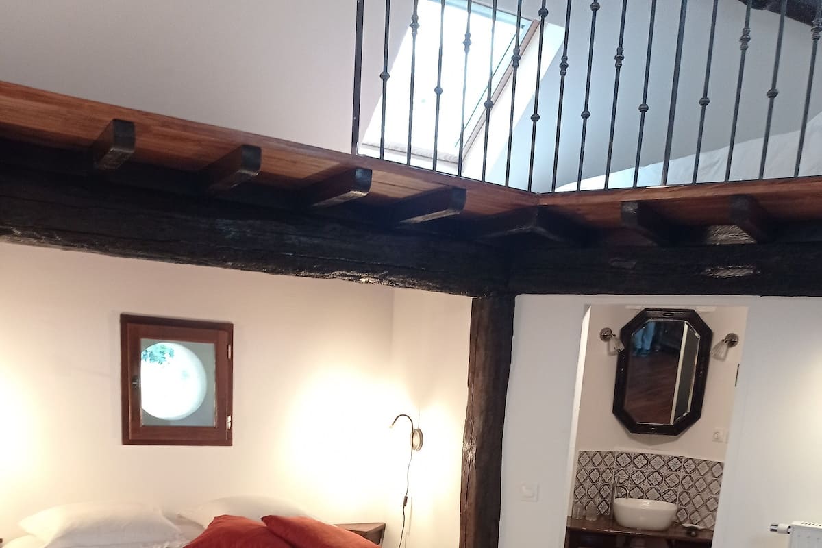 Quarto com teto de madeira, cama com roupa de cama branca, travesseiro vermelho, espelho fixado na parede e janela com vista para o céu.