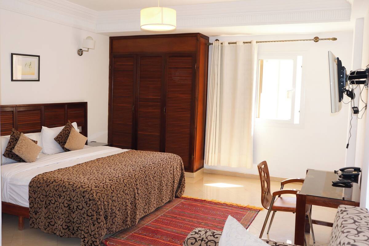 A hotel room with a large bed, a desk with a chair, a wardrobe, and a window with curtains.