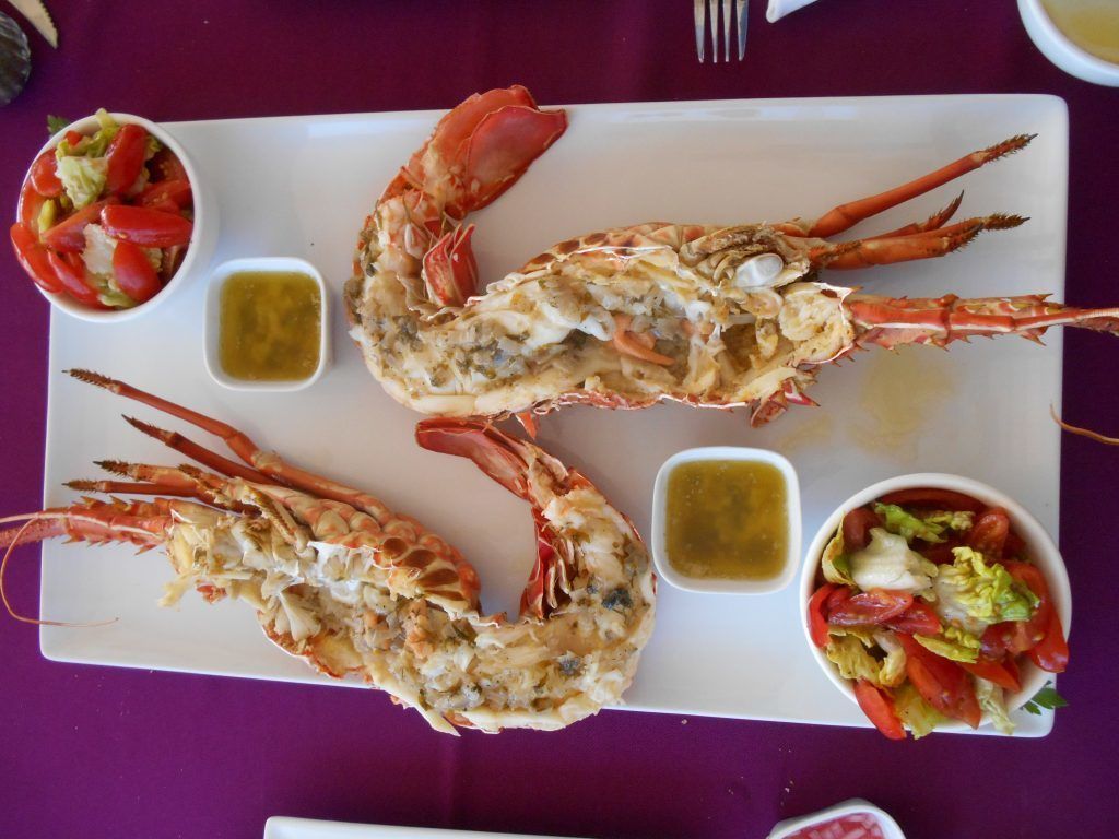 Una bandeja de mariscos con dos langostinos grandes, dos recipientes pequeños de salsa y dos acompañamientos sobre un plato rectangular blanco.