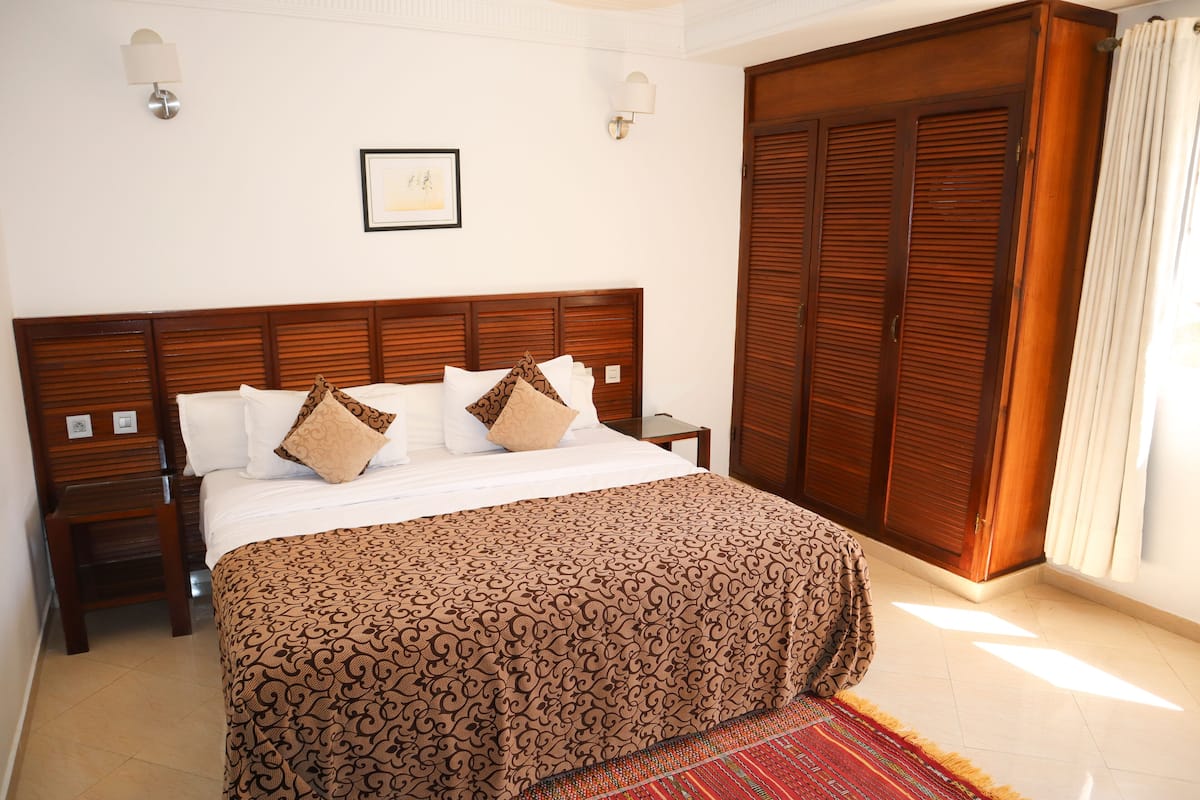 A bedroom with a large bed, wooden headboard, two bedside tables, a nightstand, a framed picture on the wall, and a large wooden wardrobe.