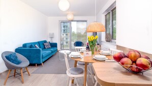 Un salon moderne avec un canapé bleu, une table à manger dressée pour deux personnes et un panier de fruits posé sur la table.