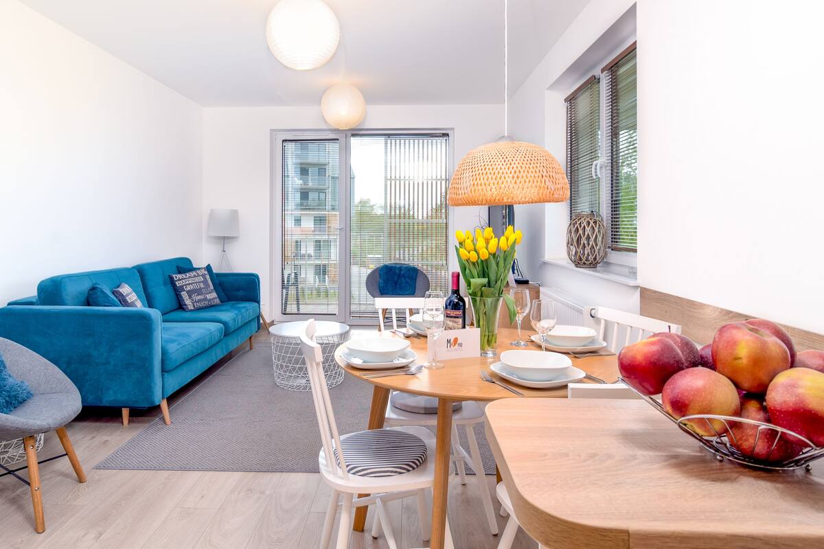 Un salon moderne avec un canapé bleu, une table à manger dressée pour deux personnes et un panier de fruits posé sur la table.